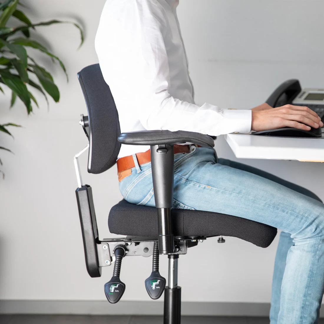 Medewerker zit rechtop op een zwarte BQE ComfortMove Office in stof aan een bureau, van opzij gezien, met de rugsteun in de lage rug, armsteunen en twee hendels onder de zitting